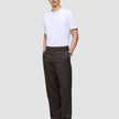 Ein Mann, der ein weißes T-Shirt und eine Atelier Pants Relaxed in Dunkelbraun aus schwerem Broken-Twill trägt und die Hände in den Taschen hat.