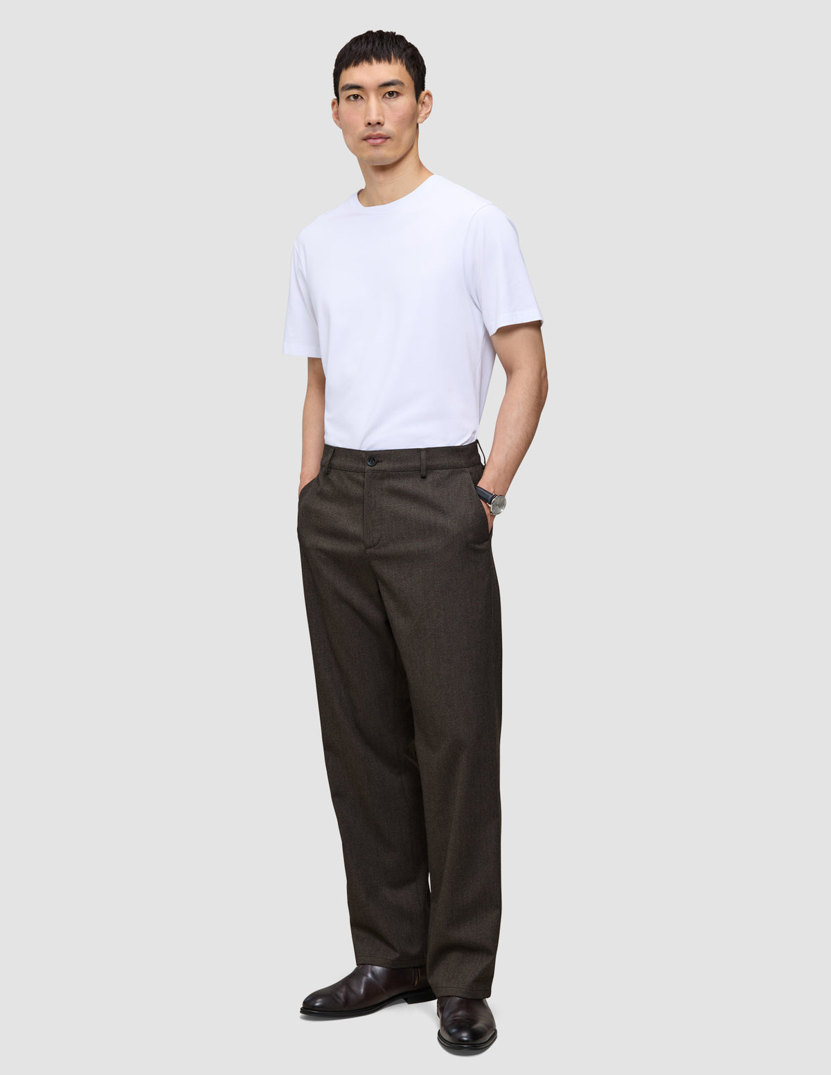 Ein Mann, der ein weißes T-Shirt und eine Atelier Pants Relaxed in Dunkelbraun aus schwerem Broken-Twill trägt und die Hände in den Taschen hat.