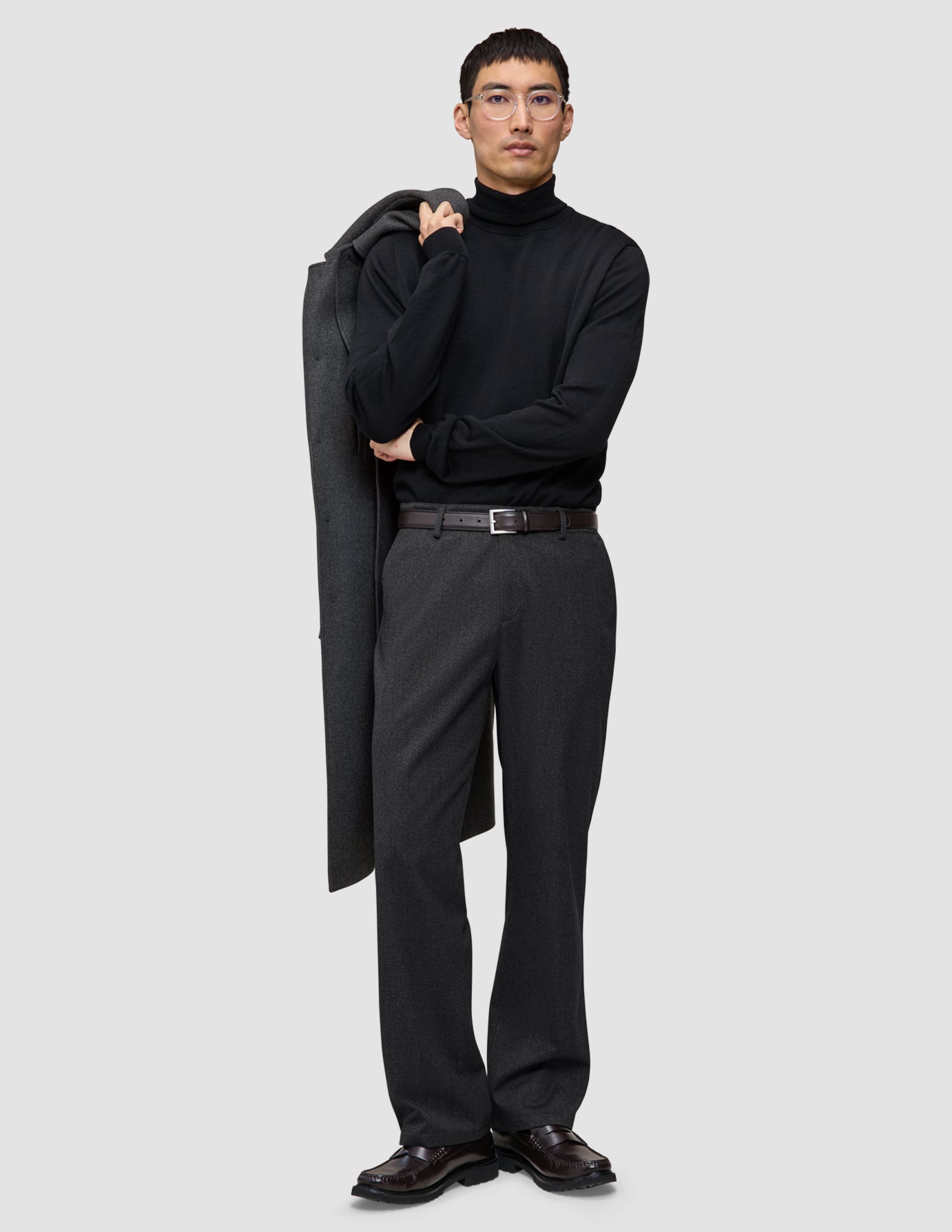 Ein Mann, der einen schwarzen Rollkragenpullover und eine Atelier Pants Relaxed in Dunkelgrau trägt und einen grauen Mantel über die Schulter gehängt hat.