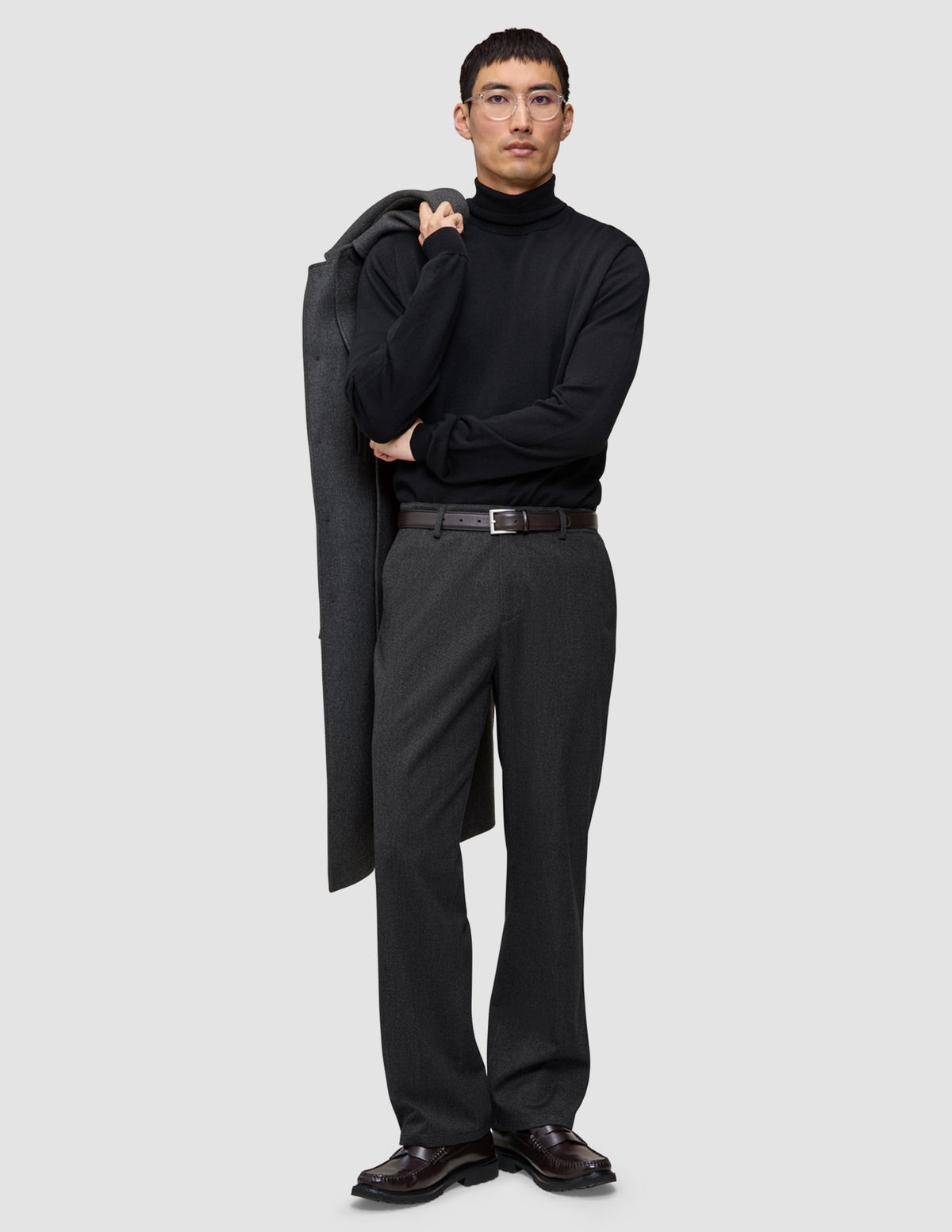 Ein Mann, der einen schwarzen Rollkragenpullover und eine Atelier Pants Relaxed in Dunkelgrau trägt und einen grauen Mantel über die Schulter gehängt hat.