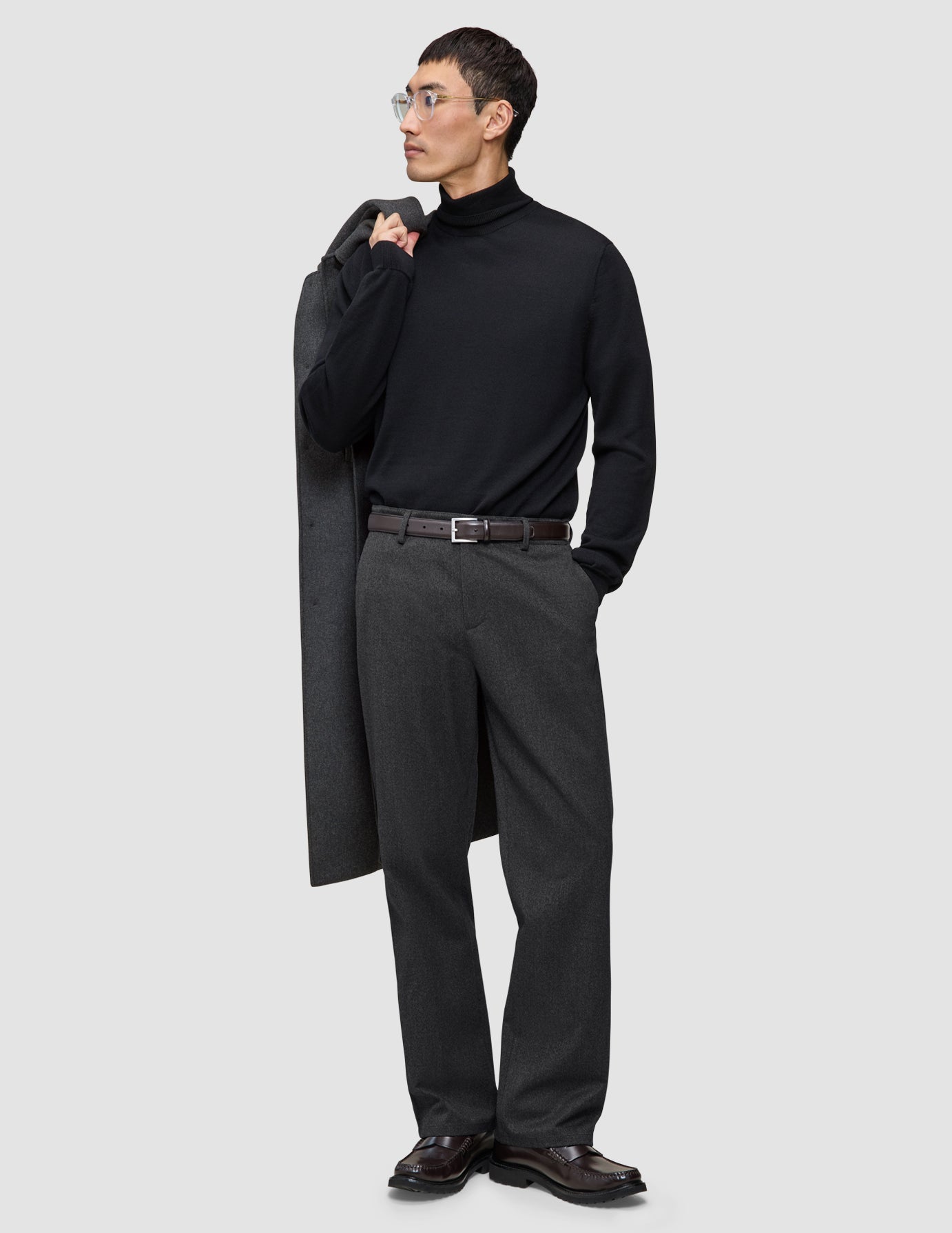 Ein Mann, der einen schwarzen Rollkragenpullover und eine Atelier Pants Relaxed in Dunkelgrau aus schwerem Broken-Twill trägt und einen Mantel über die Schulter gehängt hat.
