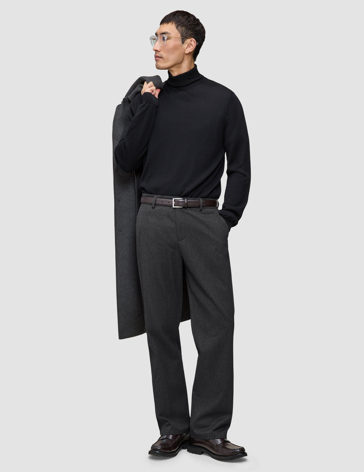 Ein Mann, der einen schwarzen Rollkragenpullover und eine Atelier Pants Relaxed in Dunkelgrau aus schwerem Broken-Twill trägt und einen Mantel über die Schulter gehängt hat.