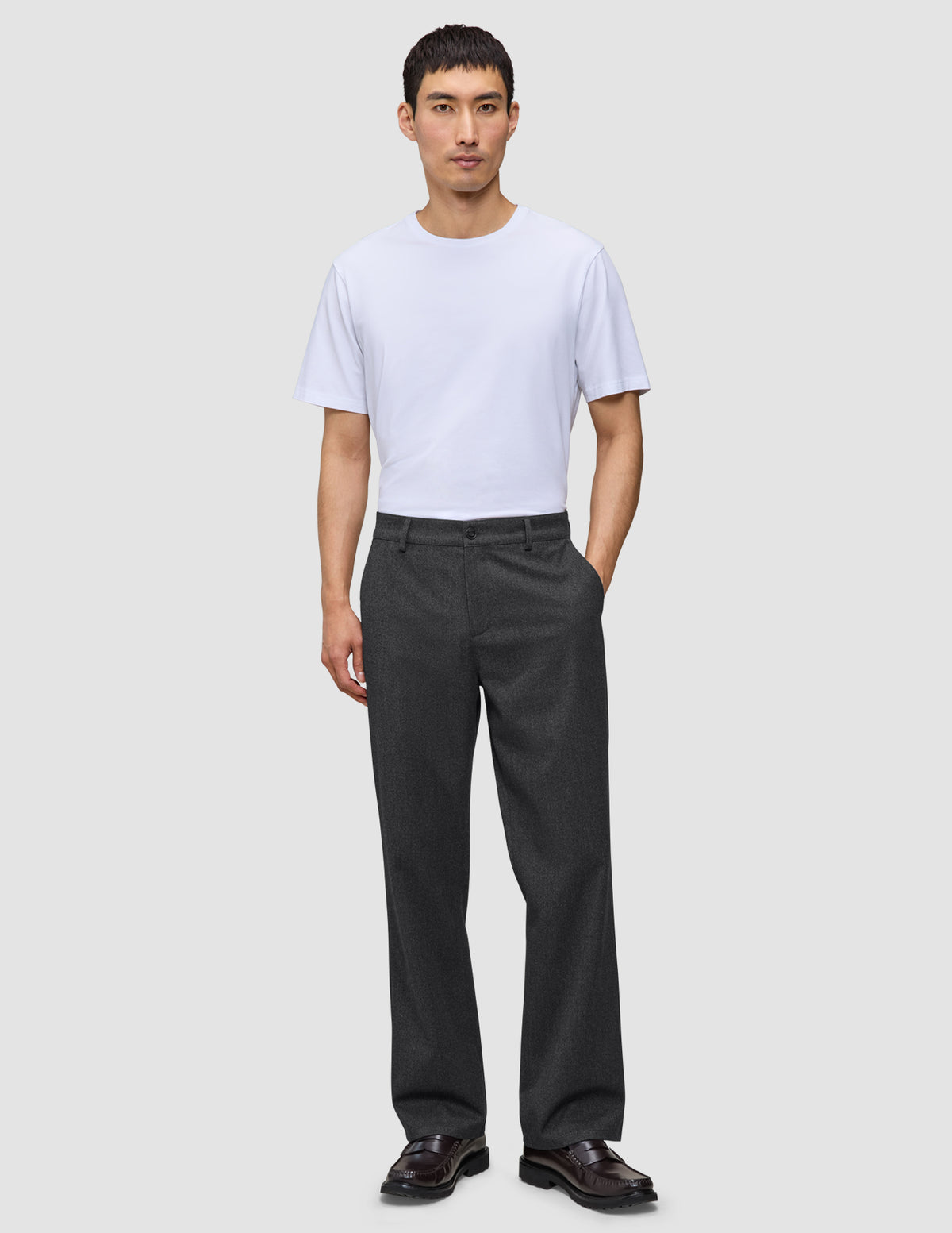 Ein Mann in einem weißen T-Shirt und einer Atelier Pants Relaxed in Dunkelgrau aus schwerem Broken-Twill, der vor einem einfarbigen Hintergrund steht.