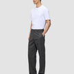 Eine Person in einem weißen T-Shirt und einer Atelier Pants Relaxed in Dunkelgrau aus schwerem Broken-Twill steht da, eine Hand in der Hosentasche.