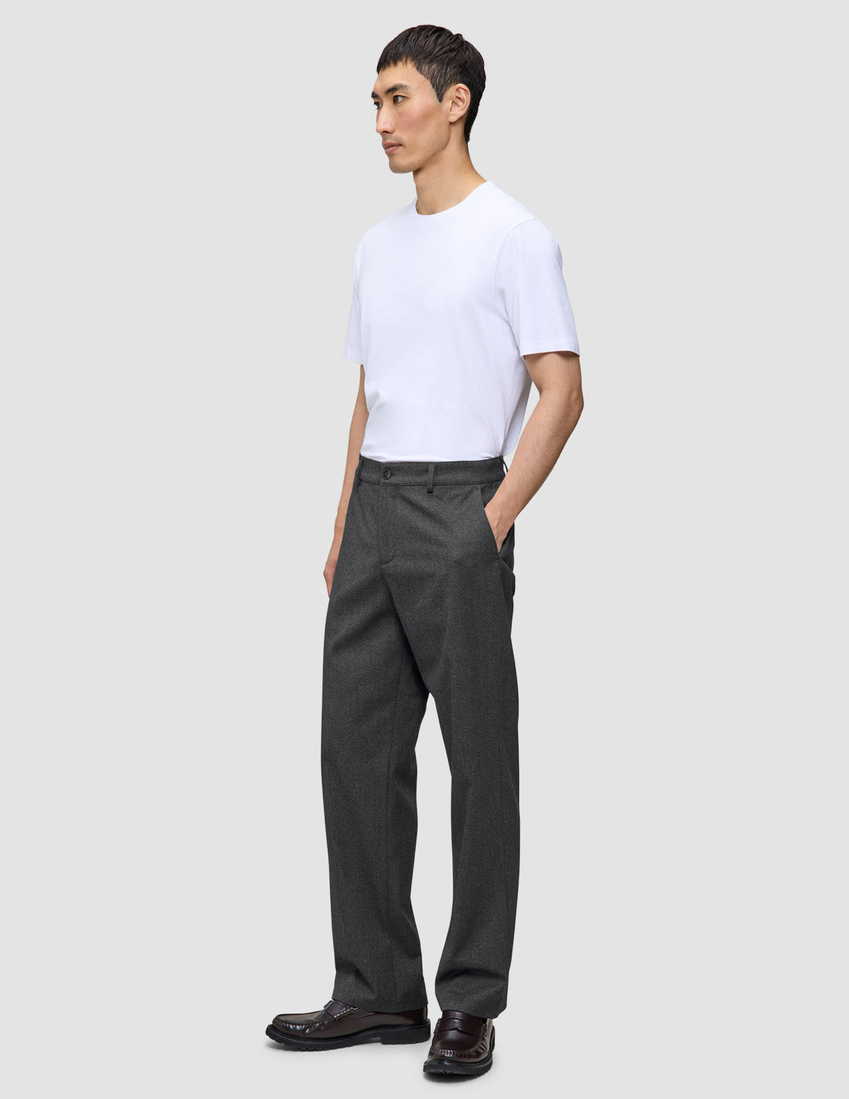 Eine Person in einem weißen T-Shirt und einer Atelier Pants Relaxed in Dunkelgrau aus schwerem Broken-Twill steht da, eine Hand in der Hosentasche.