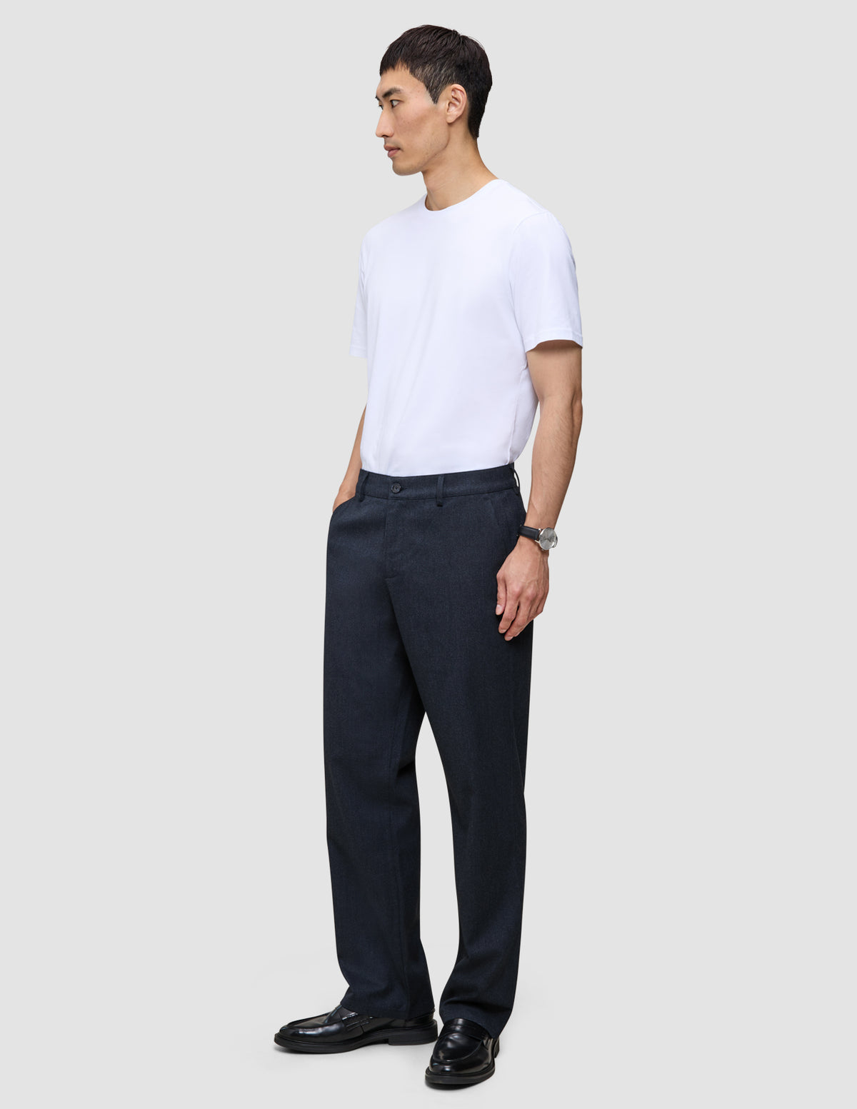 Ein Mann, der ein weißes T-Shirt und eine Atelier Pants Relaxed Dark Navy aus schwerem, gebrochenem Twill trägt, steht vor einem einfarbigen Hintergrund.