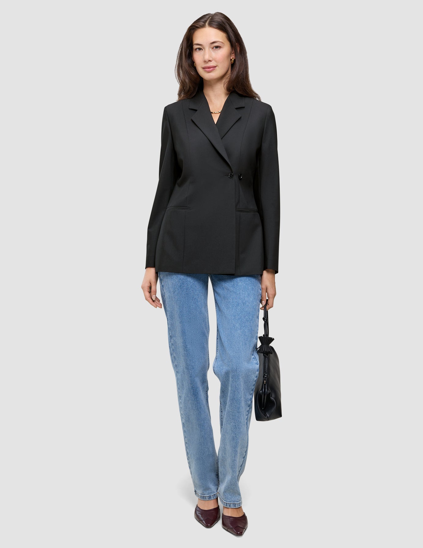 Eine Frau, die einen schwarzen, zweireihigen Women’s Avira Blazer Black-Blazer, eine blaue Jeans und dunkle Pumps trägt und eine kleine schwarze Handtasche in der Hand hält.