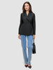 Eine Frau, die einen schwarzen, zweireihigen Women’s Avira Blazer Black-Blazer, eine blaue Jeans und dunkle Pumps trägt und eine kleine schwarze Handtasche in der Hand hält.