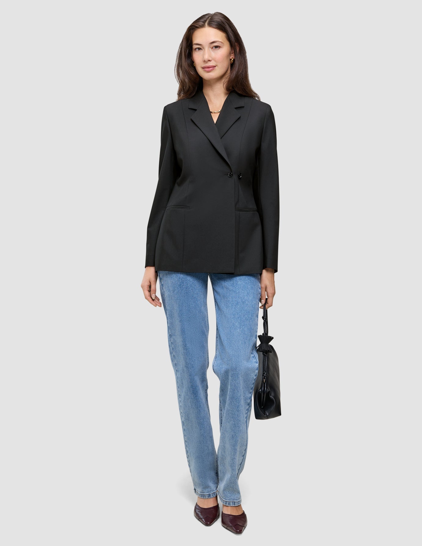 Eine Frau, die einen schwarzen, zweireihigen Women’s Avira Blazer Black-Blazer, eine blaue Jeans und dunkle Pumps trägt und eine kleine schwarze Handtasche in der Hand hält.