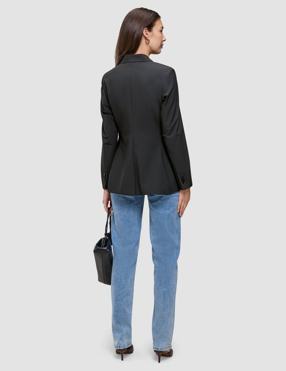 Eine Person, von hinten gesehen, die einen schwarzen Avira-Blazer für Damen, eine blaue Jeans und Pumps trägt und eine schwarze Handtasche in der Hand hält.