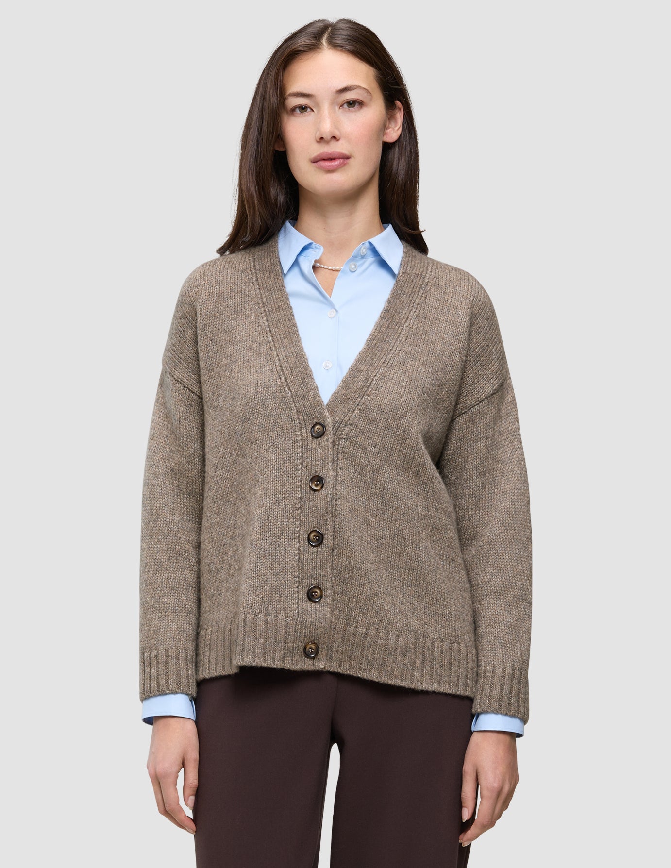 Ein Model trägt eine Damen-Strickjacke aus weicher, grobstrickiger Wolle in Taupe-Melange über einem hellblauen Hemd mit Kragen.