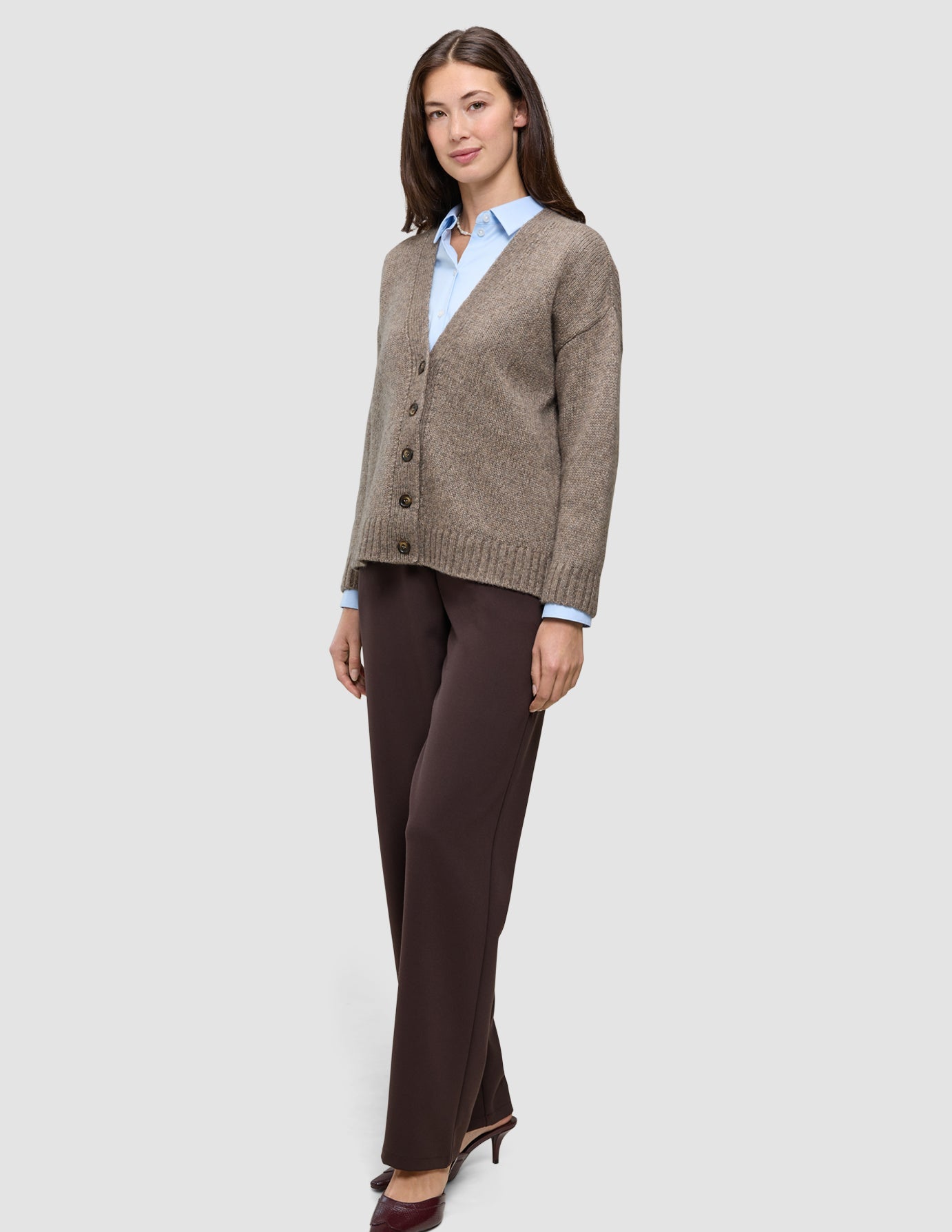 Eine Frau, die eine Damen-Strickjacke aus weicher, grobstrickiger Wolle in Taupe-Melange über einem hellblauen Hemd mit Kragen und einer dunklen Hose trägt.