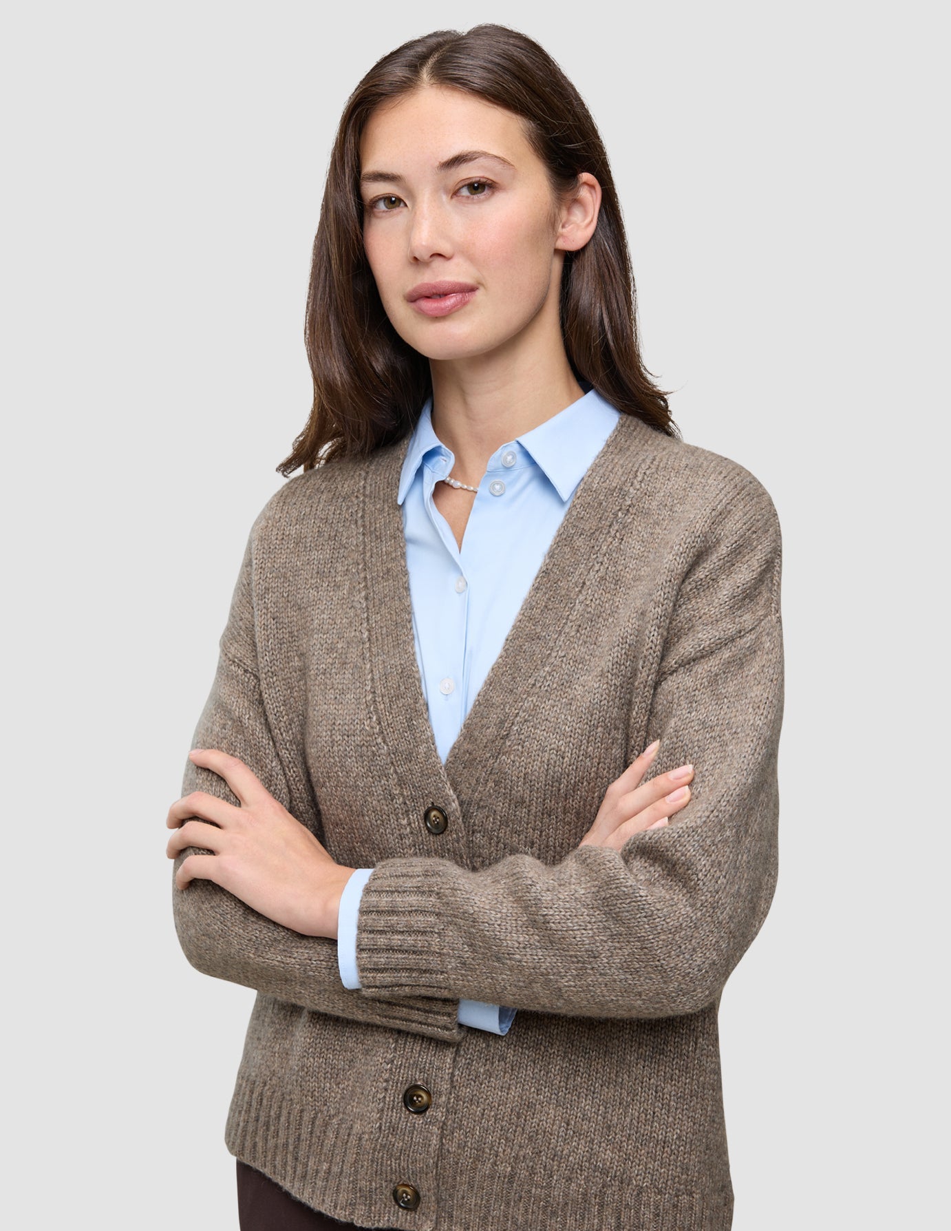 Eine Frau, die eine Damen-Strickjacke aus weicher, grobstrickiger Wolle in Taupe-Melange über einem hellblauen Hemd mit Kragen trägt.