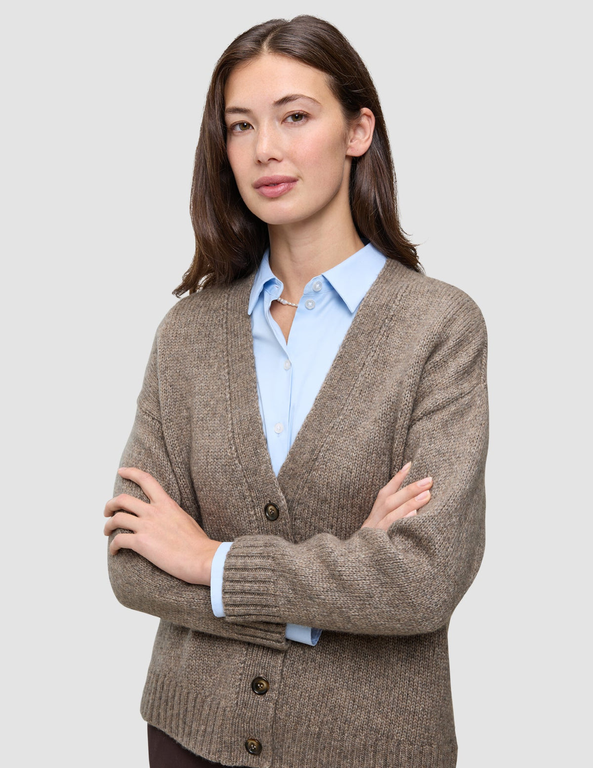 Eine Frau, die eine Damen-Strickjacke aus weicher, grobstrickiger Wolle in Taupe-Melange über einem hellblauen Hemd mit Kragen trägt.