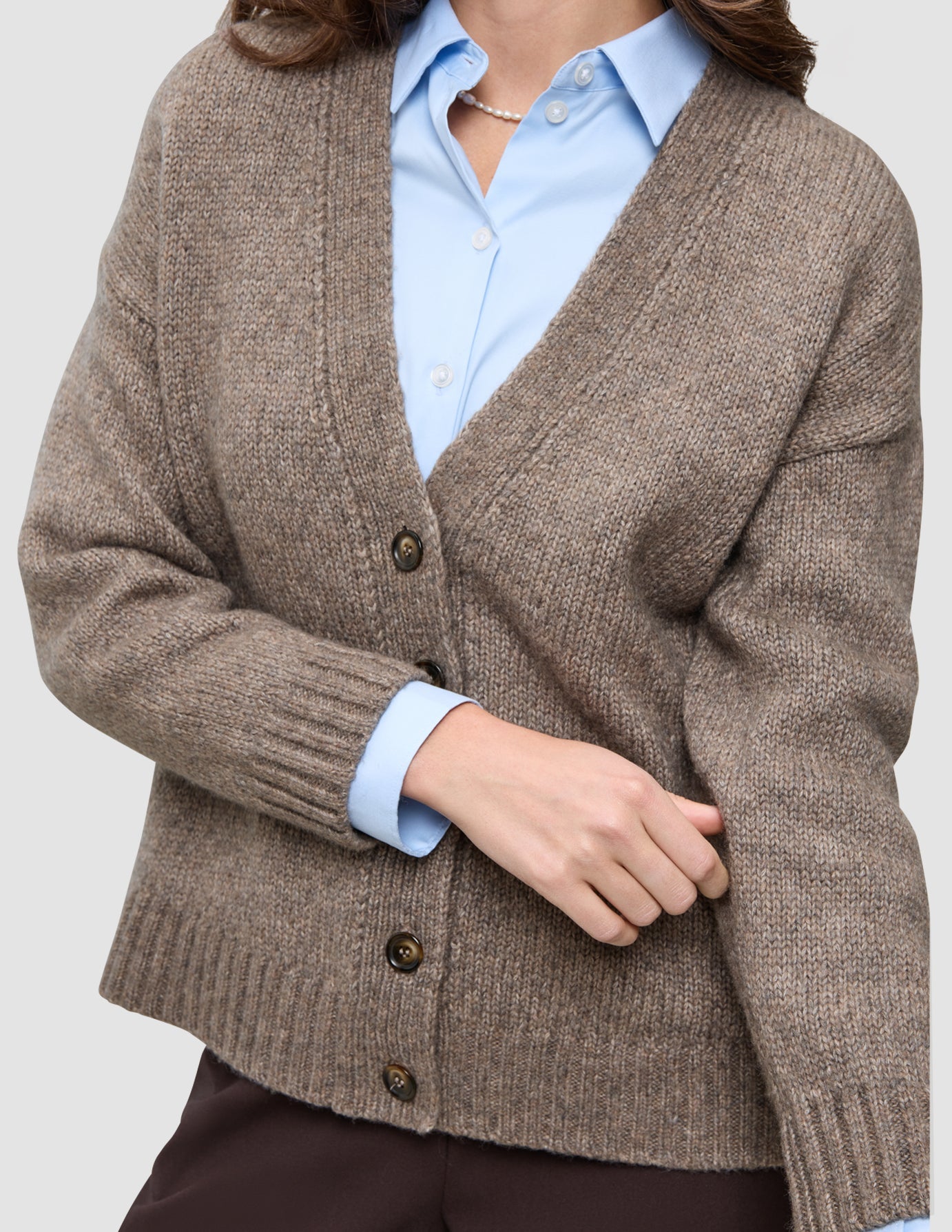 Eine grob gestrickte, weiche Wollstrickjacke für Damen in Taupe-Melange, über einem hellblauen Hemd mit Kragen geknöpft.