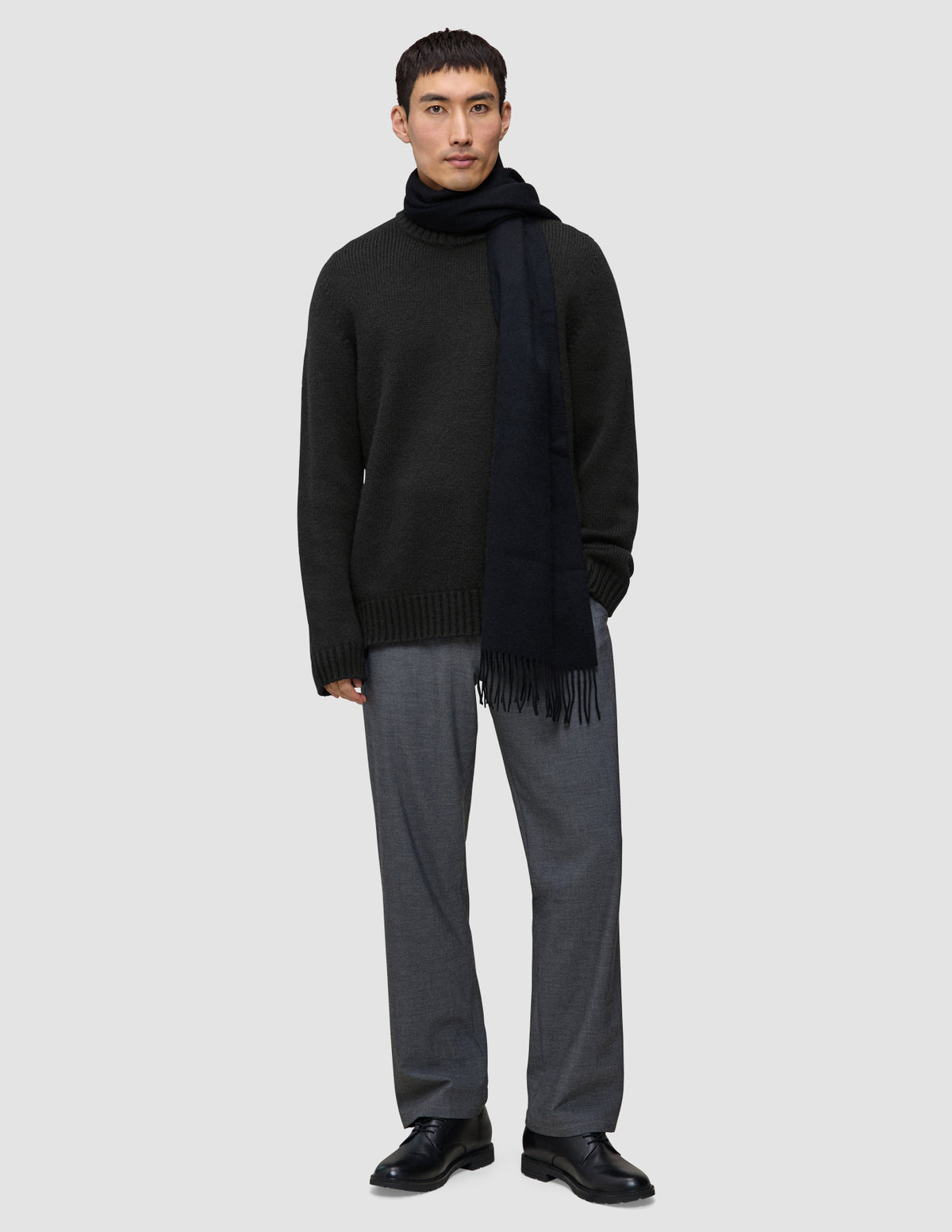 Ein Mann, der einen schwarzen, grob gestrickten, weichen Wollpullover mit Rundhalsausschnitt, einen dazu passenden Schal und eine graue Hose trägt.