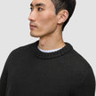 Ein Mann im Profil trägt einen schwarzen, grob gestrickten Rundhalspullover aus weicher Wolle über einem weißen T-Shirt.