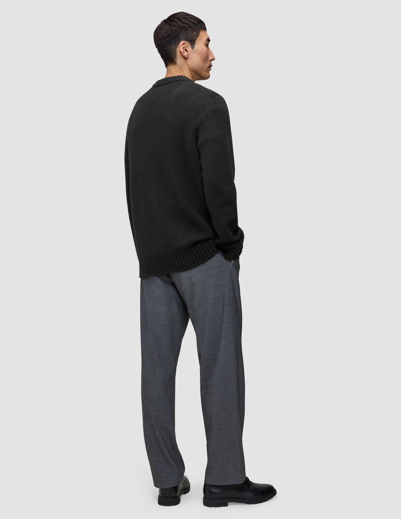 Eine Person im Profil, die einen schwarzen, grob gestrickten, weichen Wollpullover mit Rundhalsausschnitt und eine graue Hose trägt.