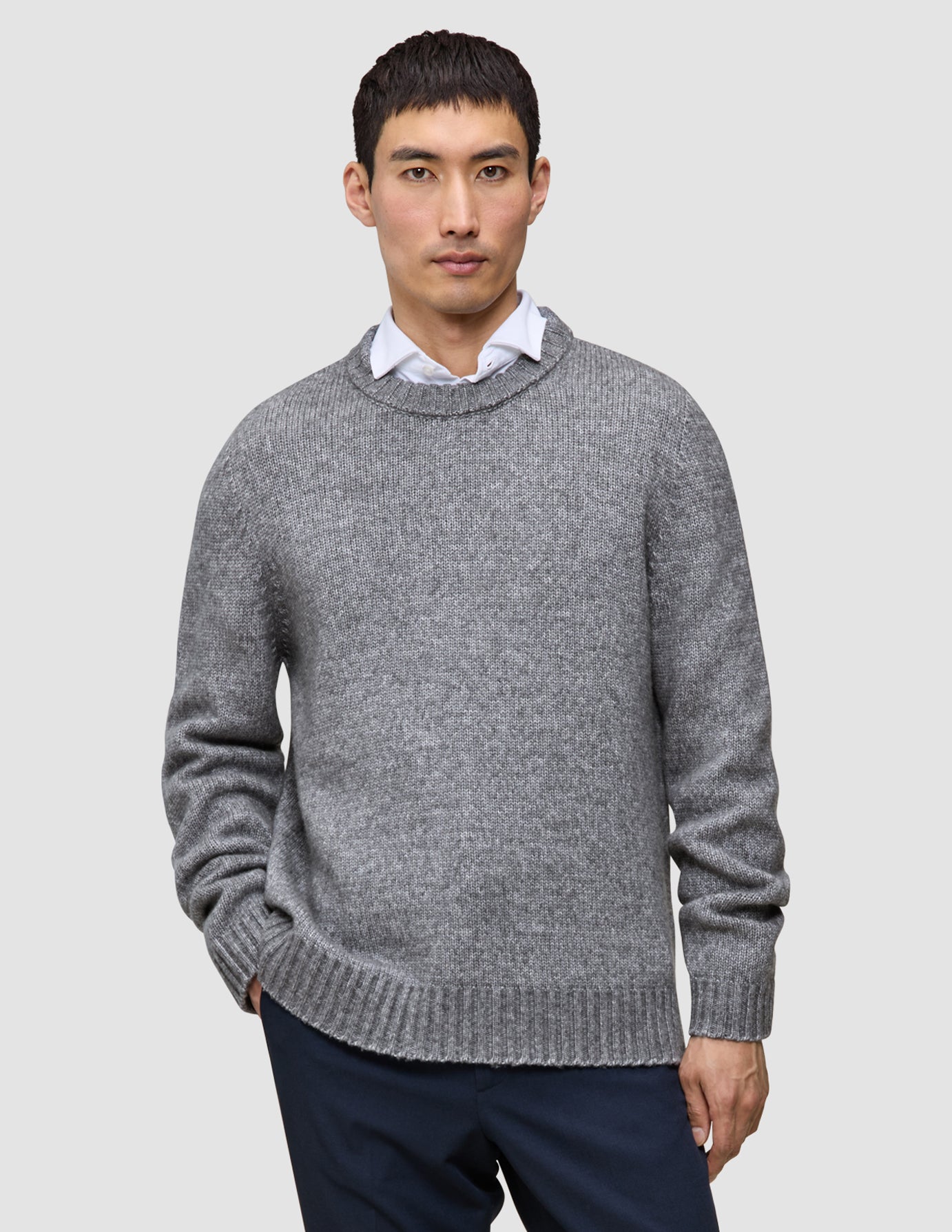 Ein Mann, der einen dicken, weichen Wollpullover mit Rundhalsausschnitt in Gletschergrau über einem weißen Hemd mit Kragen trägt.