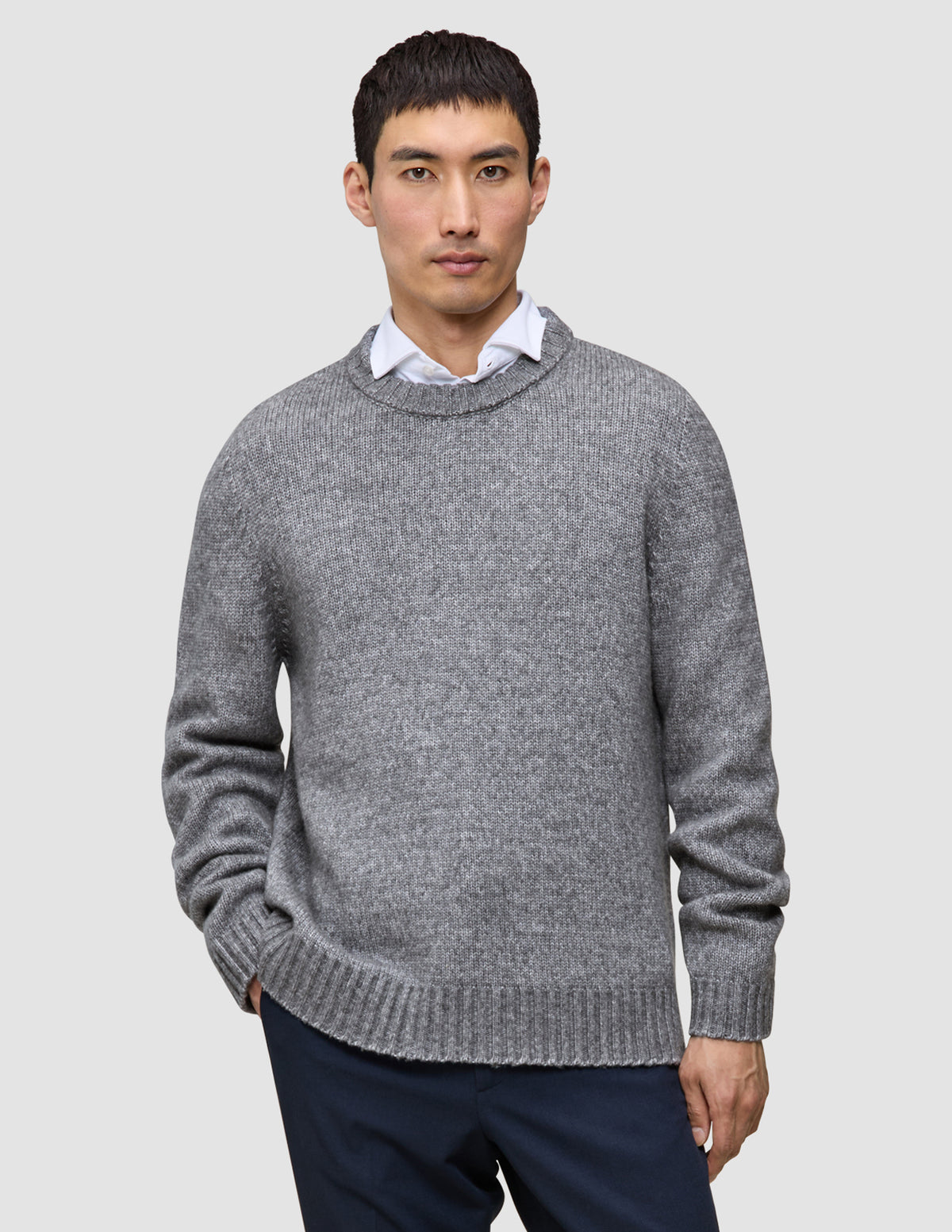 Ein Mann, der einen dicken, weichen Wollpullover mit Rundhalsausschnitt in Gletschergrau über einem weißen Hemd mit Kragen trägt.