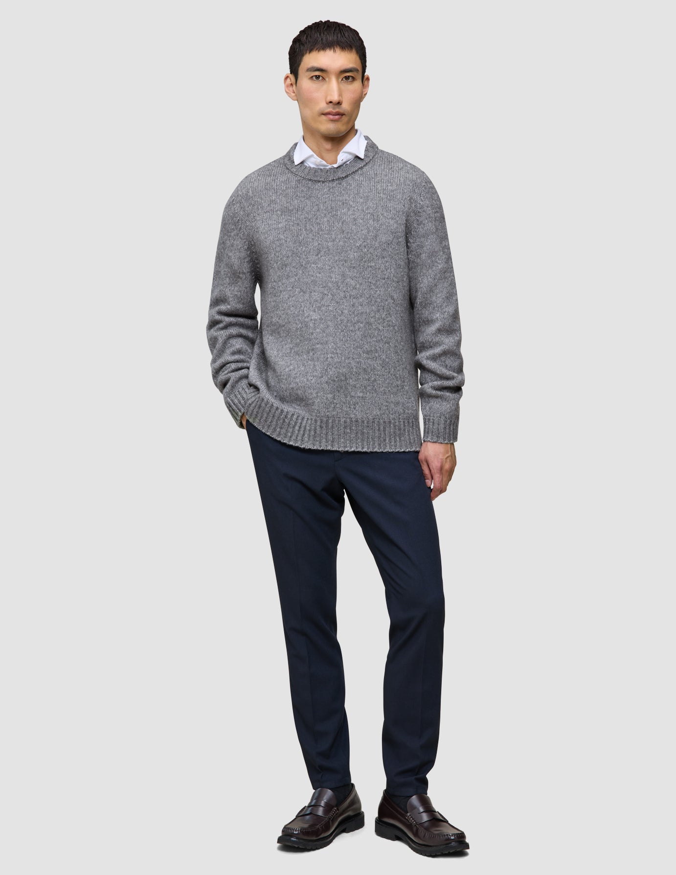 Ein Mann, der einen dicken, weichen Wollpullover mit Rundhalsausschnitt in Gletschergrau über einem weißen Hemd mit Kragen, einer dunklen Hose und braunen Slippern trägt.