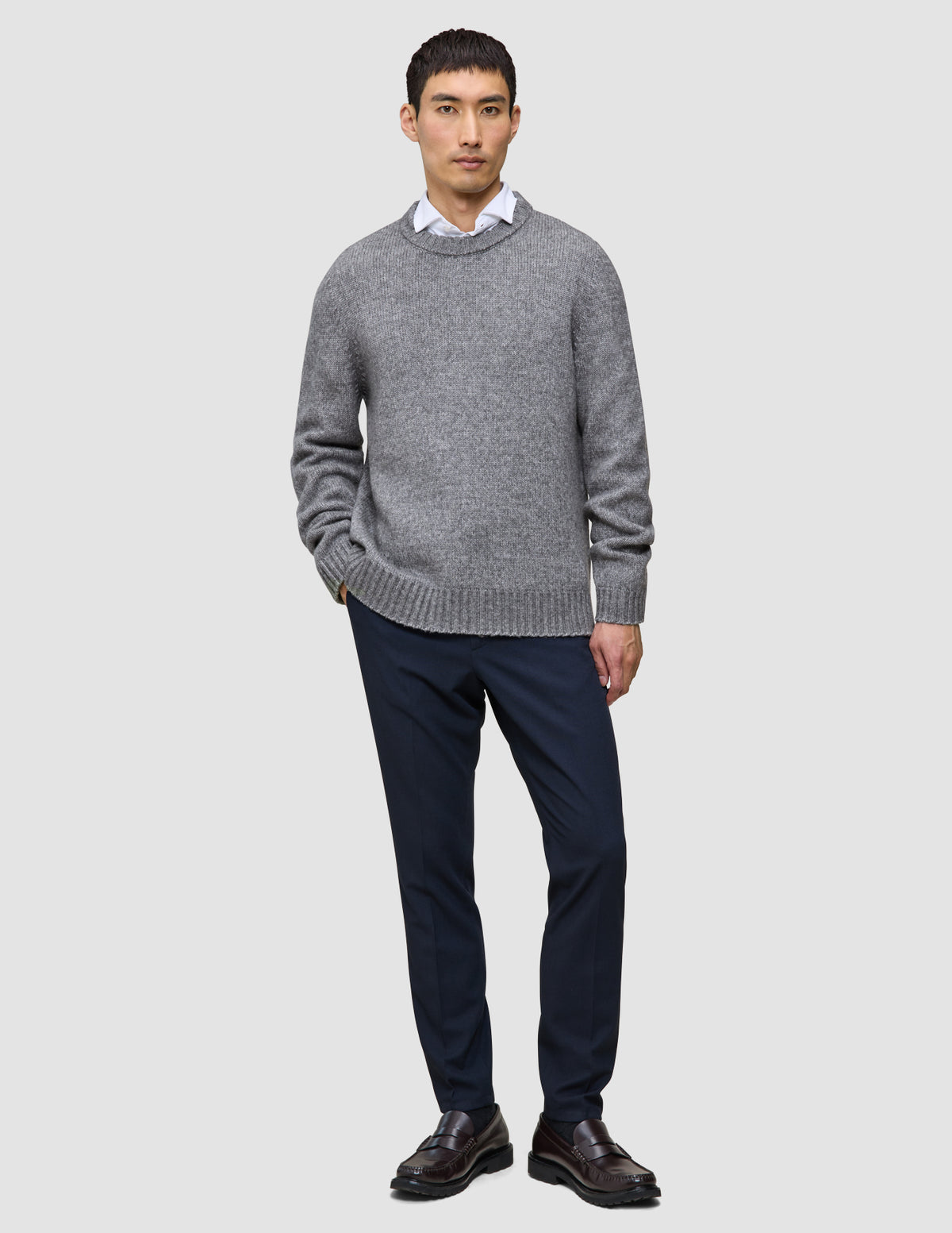 Ein Mann, der einen dicken, weichen Wollpullover mit Rundhalsausschnitt in Gletschergrau über einem weißen Hemd mit Kragen, einer dunklen Hose und braunen Slippern trägt.