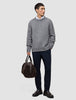 Ein Mann in einem dicken, weichen Wollpullover mit Rundhalsausschnitt in Gletschergrau, den er über einem weißen Hemd mit Kragen trägt, dazu eine marineblaue Hose und dunkle Slipper; in der Hand hält er eine braune Reisetasche.