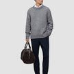 Ein Mann in einem dicken, weichen Wollpullover mit Rundhalsausschnitt in Gletschergrau, den er über einem weißen Hemd mit Kragen trägt, dazu eine marineblaue Hose und dunkle Slipper; in der Hand hält er eine braune Reisetasche.