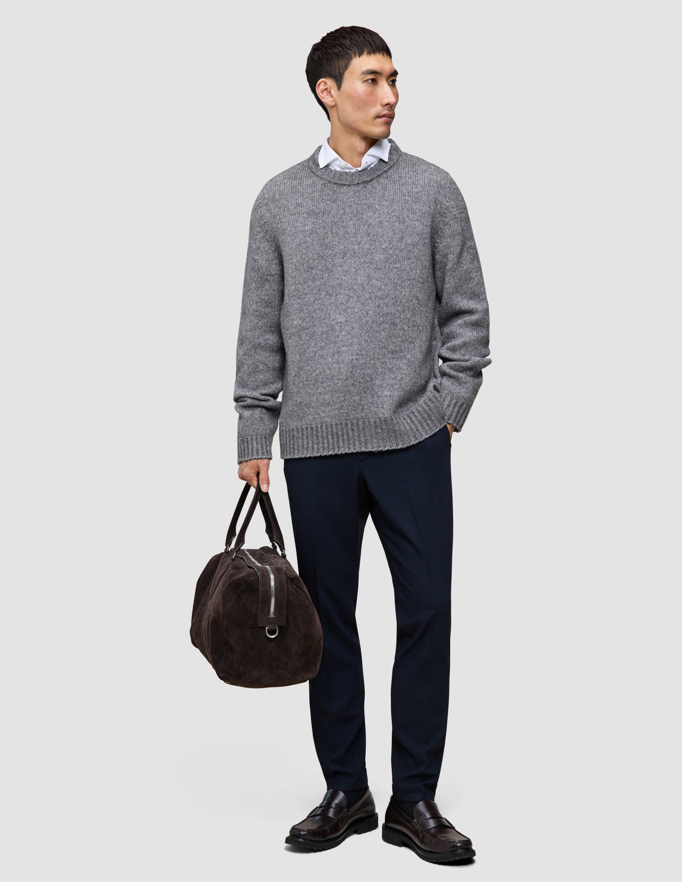 Ein Mann in einem dicken, weichen Wollpullover mit Rundhalsausschnitt in Gletschergrau, den er über einem weißen Hemd mit Kragen trägt, dazu eine marineblaue Hose und dunkle Slipper; in der Hand hält er eine braune Reisetasche.