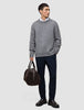 Ein Mann trägt einen dicken, weichen Wollpullover mit Rundhalsausschnitt in Gletschergrau über einem weißen Hemd mit Kragen und einer dunklen Hose und hält eine braune Reisetasche in der Hand.