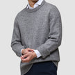 Ein Mann trägt einen dicken, weichen Wollpullover mit Rundhalsausschnitt in der Farbe Glacier Grey über einem weißen Hemd mit Kragen.