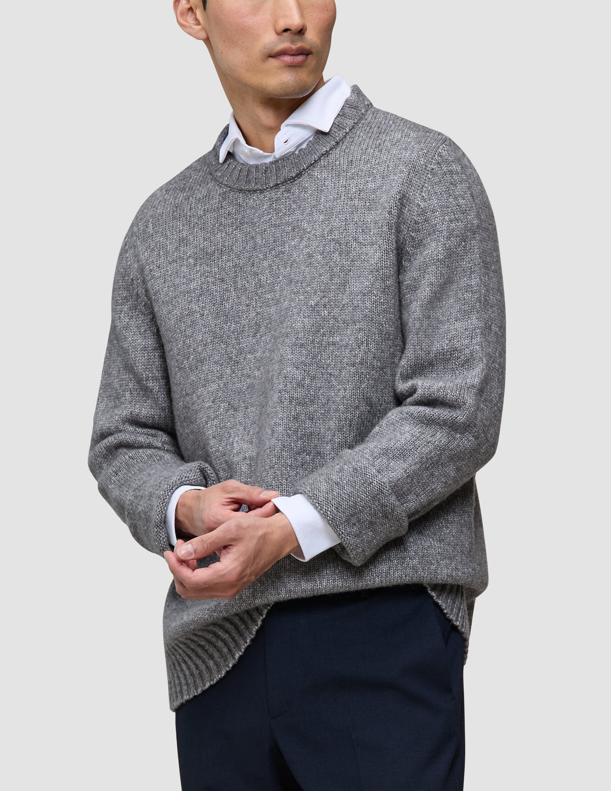Ein Mann, der einen dicken, weichen Wollpullover mit Rundhalsausschnitt in der Farbe Glacier Grey über einem weißen Hemd mit Kragen und einer dunklen Hose trägt.