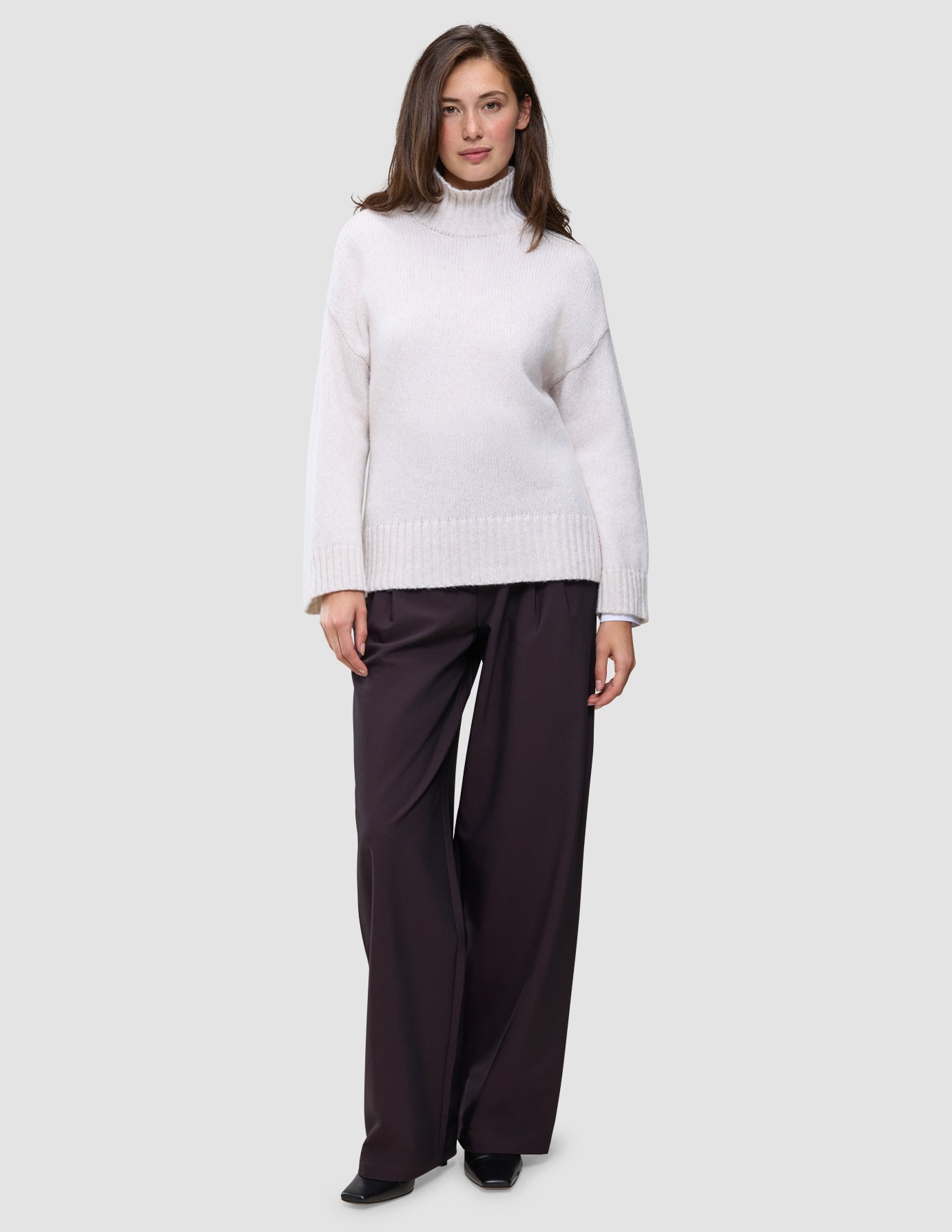 Eine Frau, die einen Damen-Rollkragenpullover aus dicker, weicher Wolle in der Farbe Sugar Melange in Off-White und eine dunkle Hose mit weitem Bein trägt.