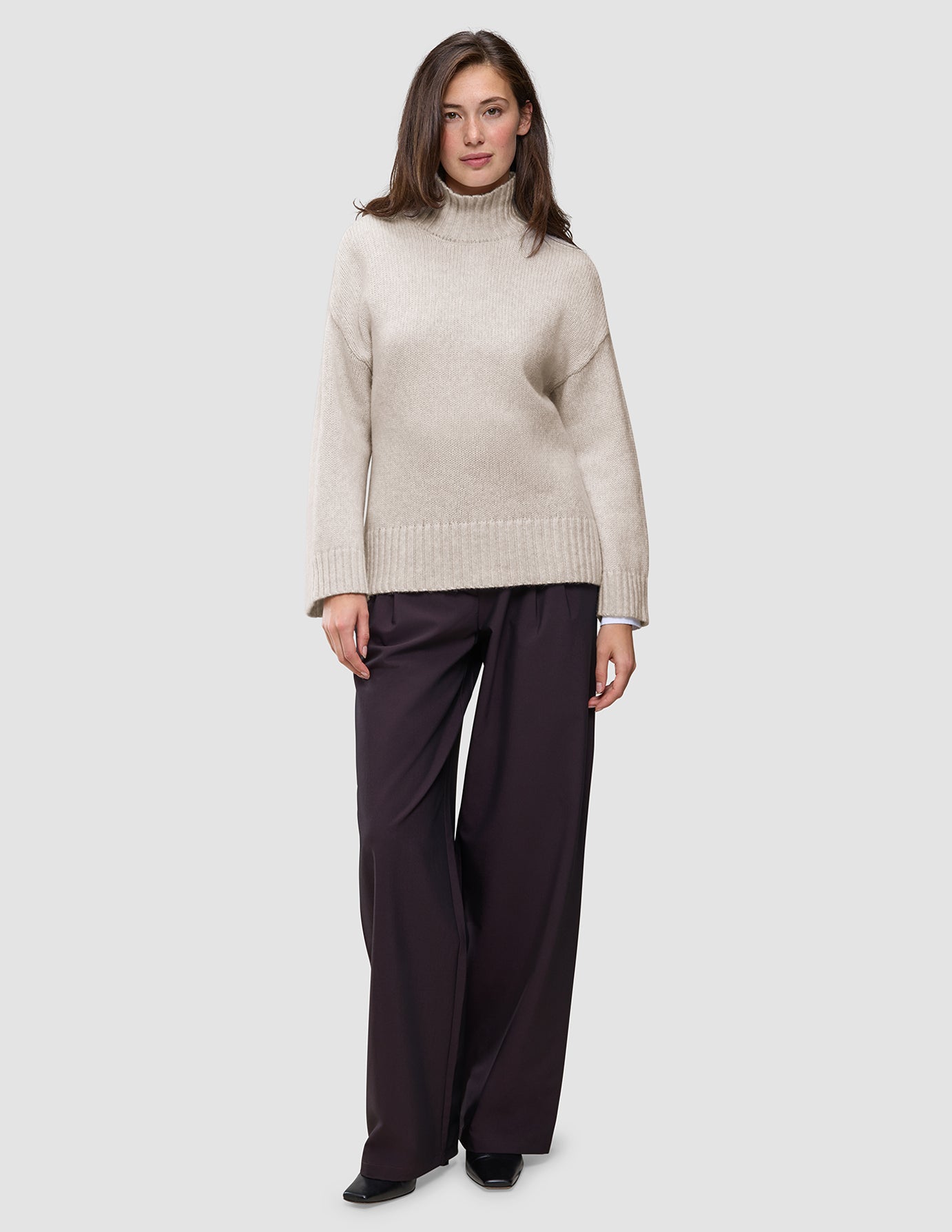 Eine Frau trägt einen übergroßen, beigen Damen-Rollkragenpullover aus weicher, grobstrickiger Wolle in der Farbe Sugar Melange mit langen Ärmeln und eine dunkle Hose mit weitem Bein.