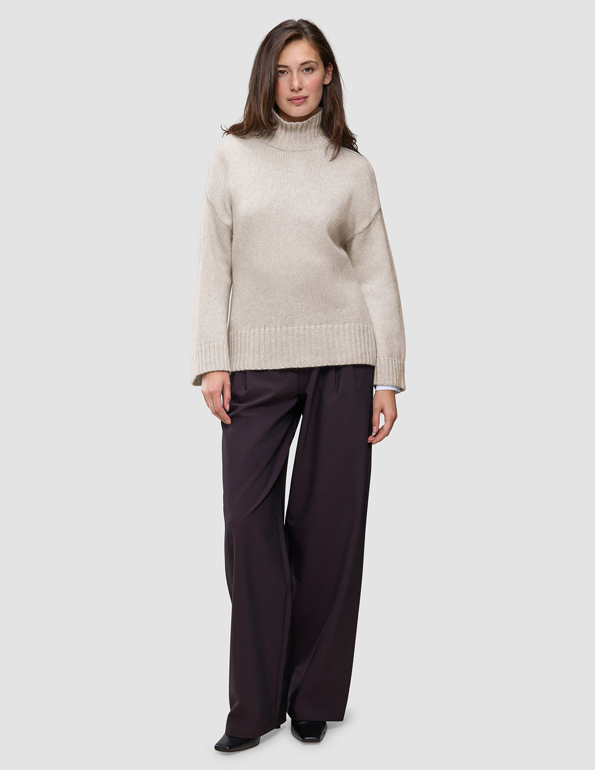 Eine Frau trägt einen übergroßen, beigen Damen-Rollkragenpullover aus weicher, grobstrickiger Wolle in der Farbe Sugar Melange mit langen Ärmeln und eine dunkle Hose mit weitem Bein.