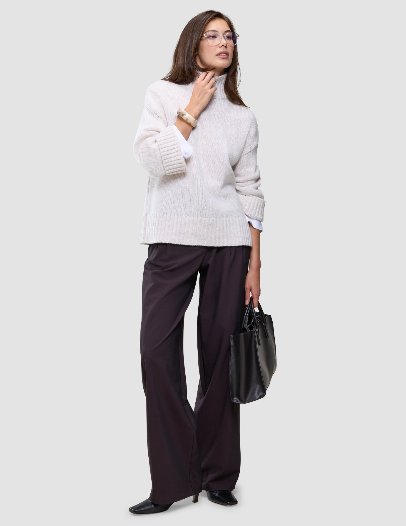 Eine Frau, die einen Damen-Rollkragenpullover aus weicher, grobstrickiger Wolle in der Farbe Sugar Melange, eine dunkle Hose mit weitem Bein und eine Brille trägt und eine schwarze Handtasche in der Hand hält.