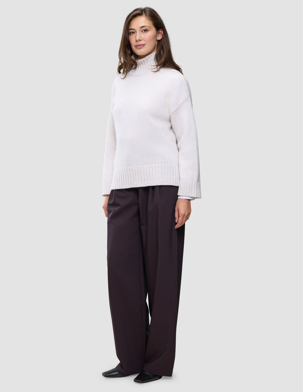 Eine Frau trägt einen cremefarbenen Damen-Rollkragenpullover aus dickem, weichem Wollstrick in der Farbe Sugar Melange sowie eine dunkle Hose mit weitem Bein.