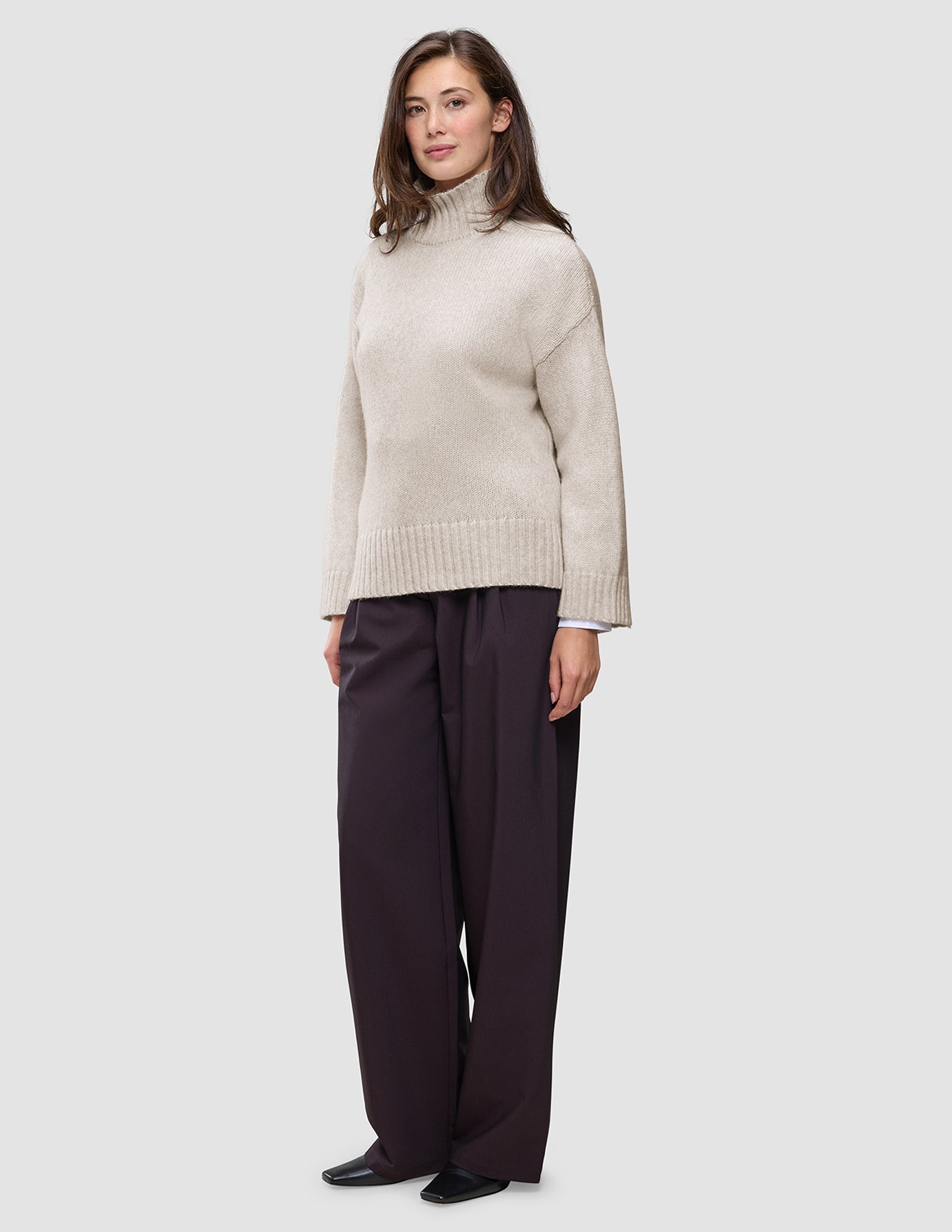 Eine Frau, die einen beigen Damen-Rollkragenpullover aus dickem, weichem Wollstrick in der Farbe Sugar Melange und eine dunkle Hose mit weitem Bein trägt.