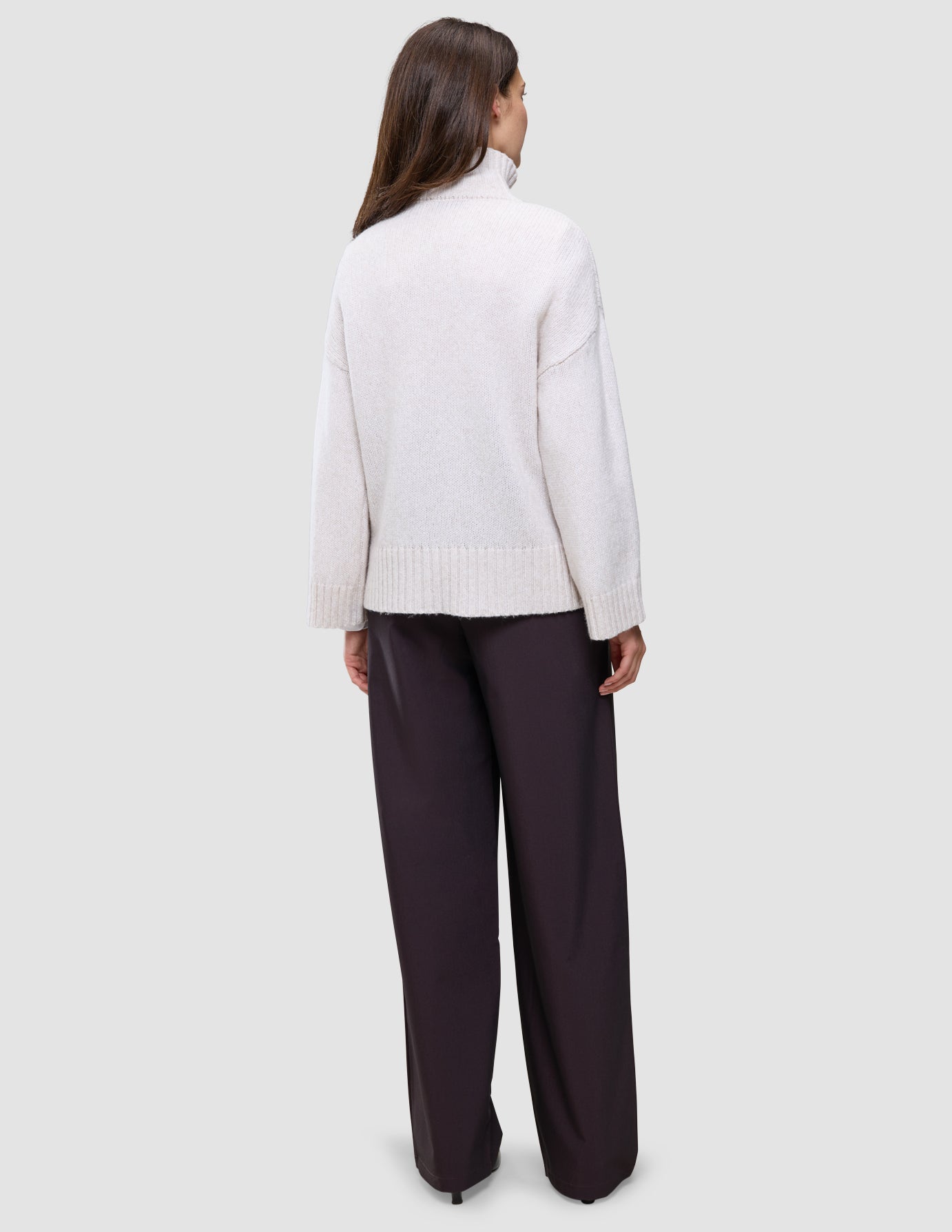 Eine Person, von hinten gesehen, die einen Damen-Rollkragenpullover aus weicher, grobstrickiger Wolle in der Farbe Sugar Melange sowie eine dunkle Hose mit weitem Bein trägt.