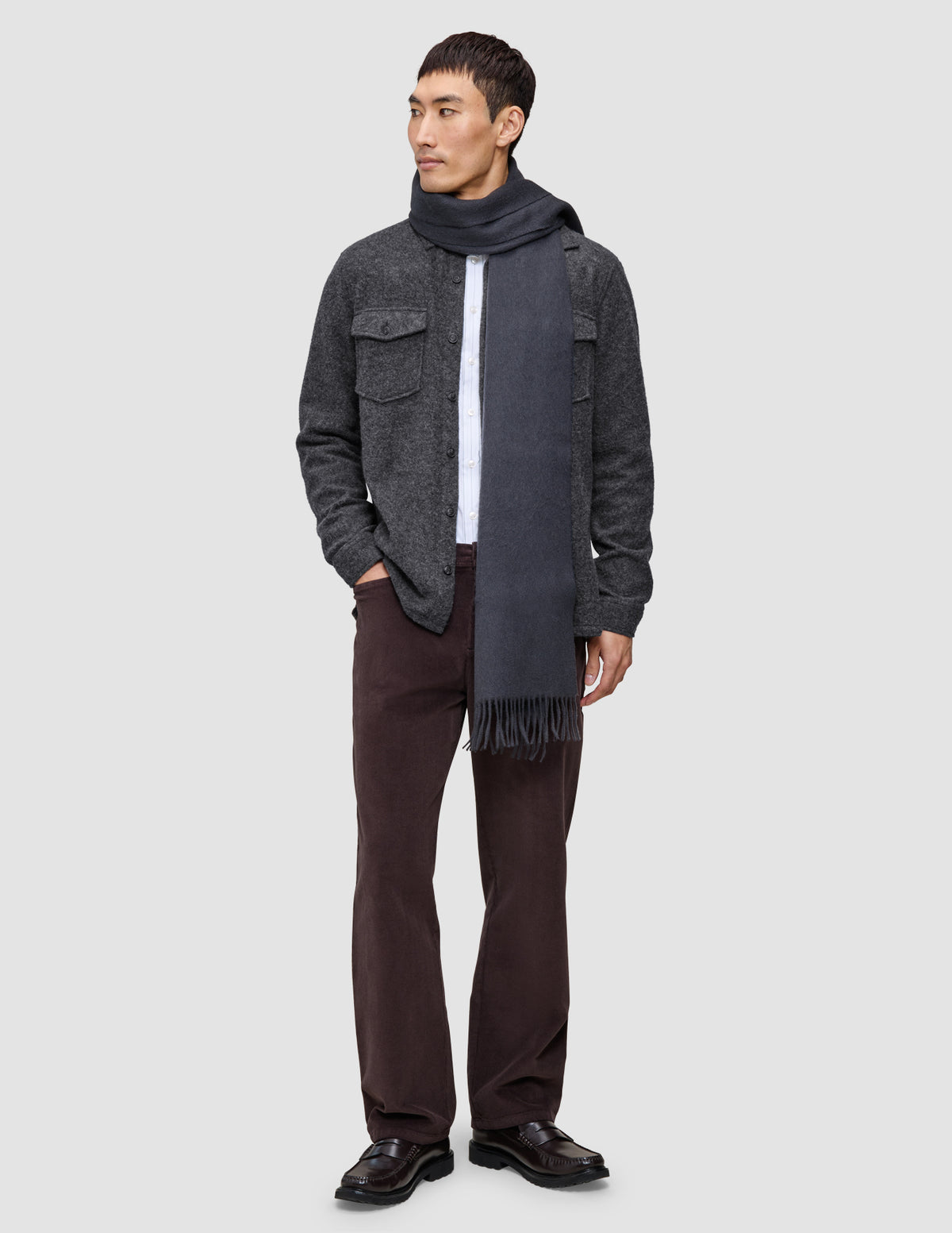 Ein Mann, der ein doppellagiges Woll-Overshirt in Regular-Passform und Dunkelgrau über einem weißen Hemd trägt, dazu eine dunkle Hose, dunkle Slipper und einen langen grauen Schal.