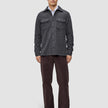 Ein Mann trägt ein doppellagiges Woll-Overshirt in Regular-Passform in Dunkelgrau über einem weißen Hemd, dazu eine dunkle Hose und schwarze Loafer im skandinavischen Design.