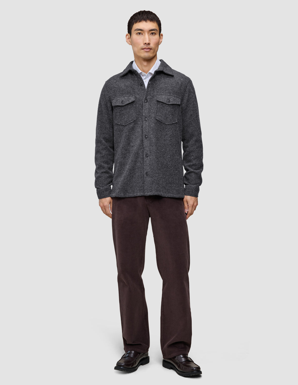 Ein Mann trägt ein doppellagiges Woll-Overshirt in Regular-Passform in Dunkelgrau über einem weißen Hemd, dazu eine dunkle Hose und schwarze Loafer im skandinavischen Design.