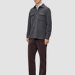 Ein Mann, der ein doppellagiges Woll-Overshirt in Regular-Passform und Dunkelgrau über einem weißen Hemd mit Kragen trägt, dazu eine dunkle Hose und braune Slipper in einem schlichten, skandinavischen Design.