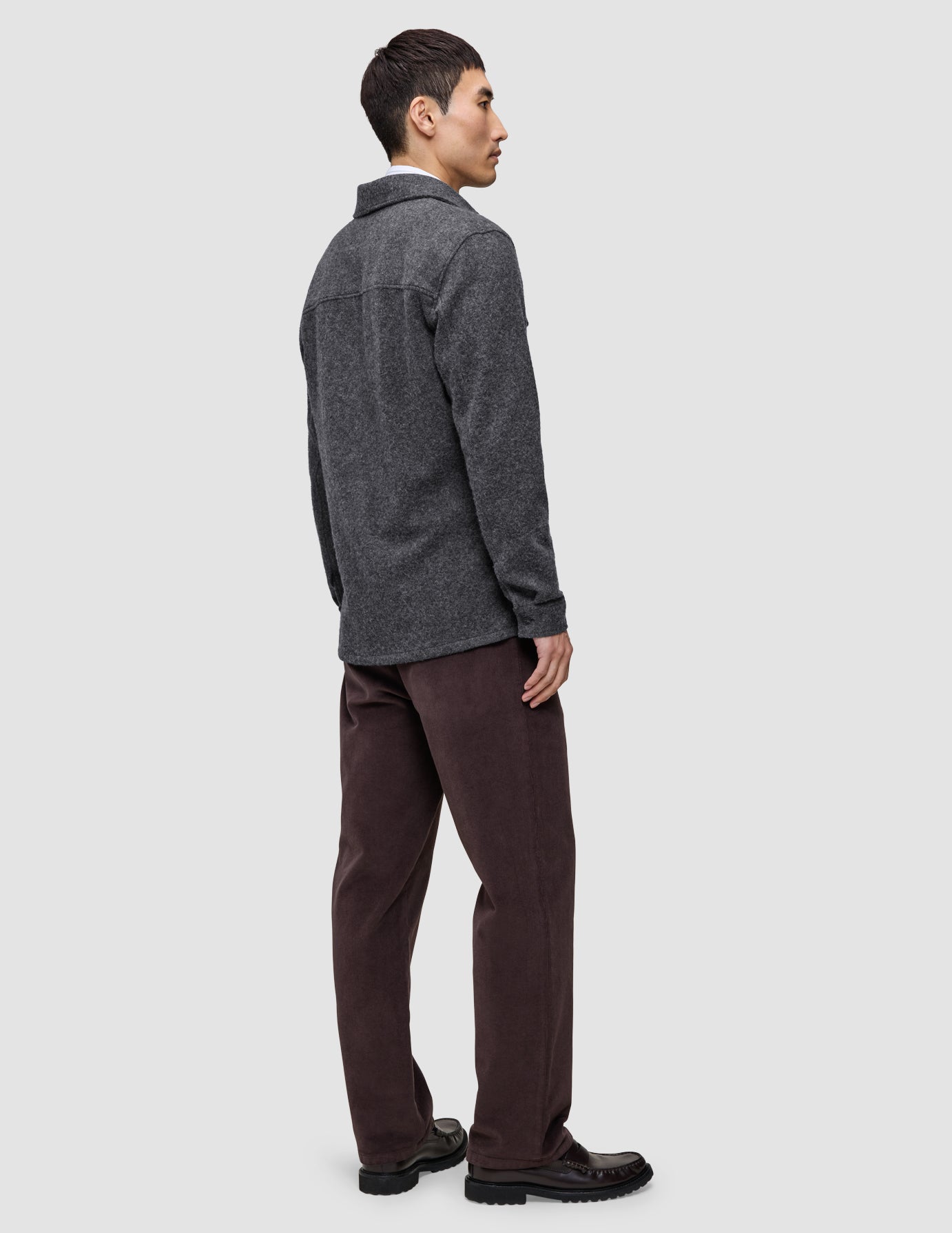 Eine Person, von hinten gesehen, die ein doppellagiges Woll-Overshirt in der Ausführung Regular in Dunkelgrau, eine dunkelbraune Hose und schwarze Slipper trägt.