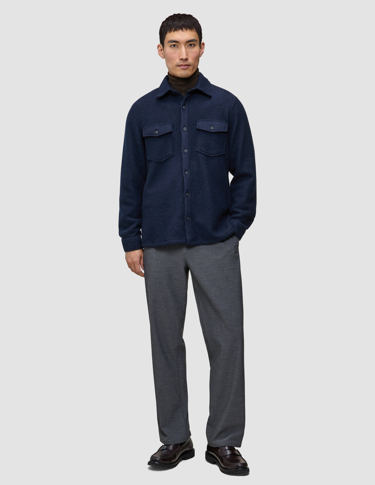 Ein Mann trägt ein doppellagiges Wollhemd in der Farbe Regular Dark Navy über einem dunklen Rollkragenpullover, dazu eine graue Hose und dunkle Leder-Loafer im minimalistischen skandinavischen Design.