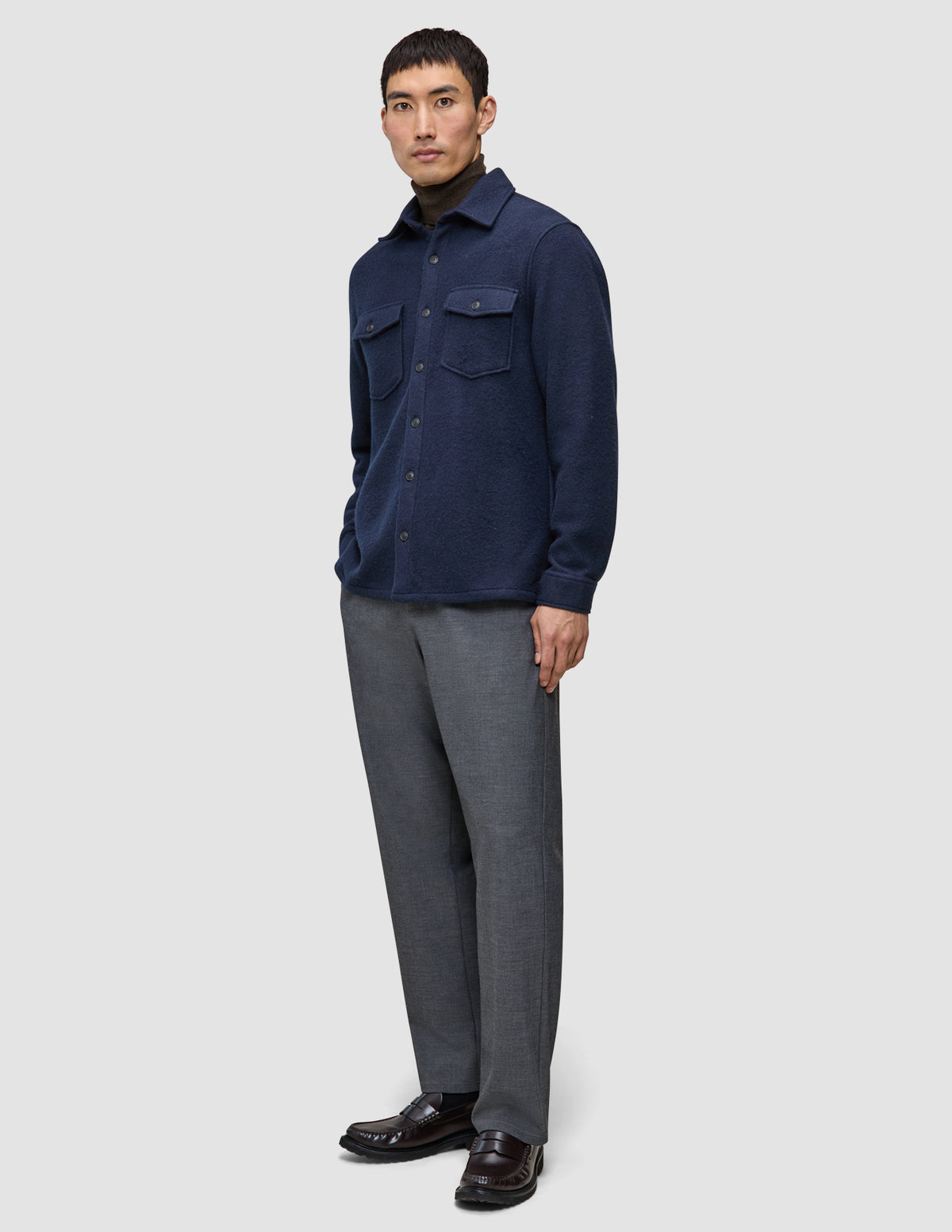 Ein Mann, der ein doppellagiges Wollhemd in der Farbe Regular Dark Navy über einem braunen Rollkragenpullover trägt, dazu eine graue Hose und dunkle Slipper.