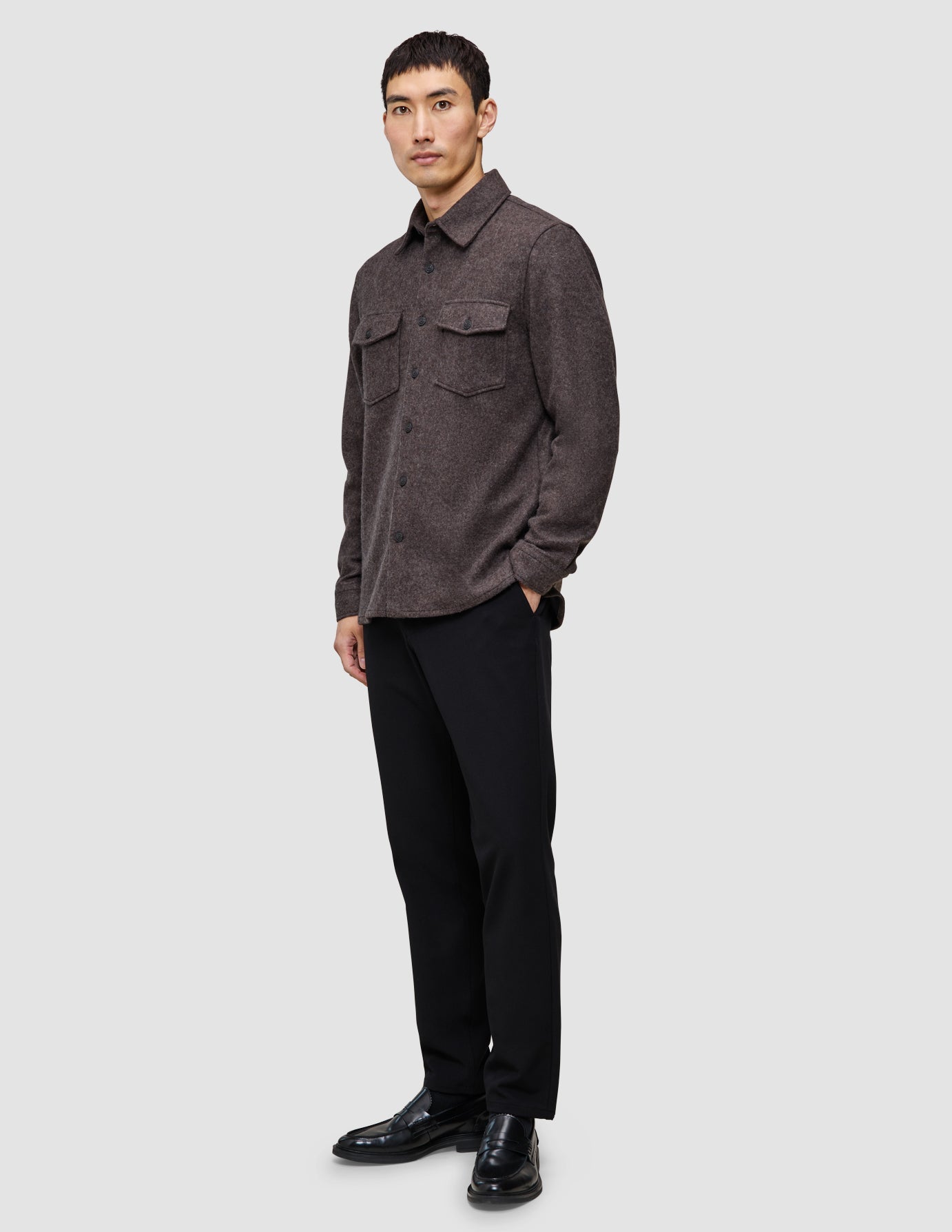 Eine Person, die ein doppellagiges Woll-Overshirt in Sattelbraun mit Brusttaschen, eine schwarze Hose und schwarze Loafer trägt.