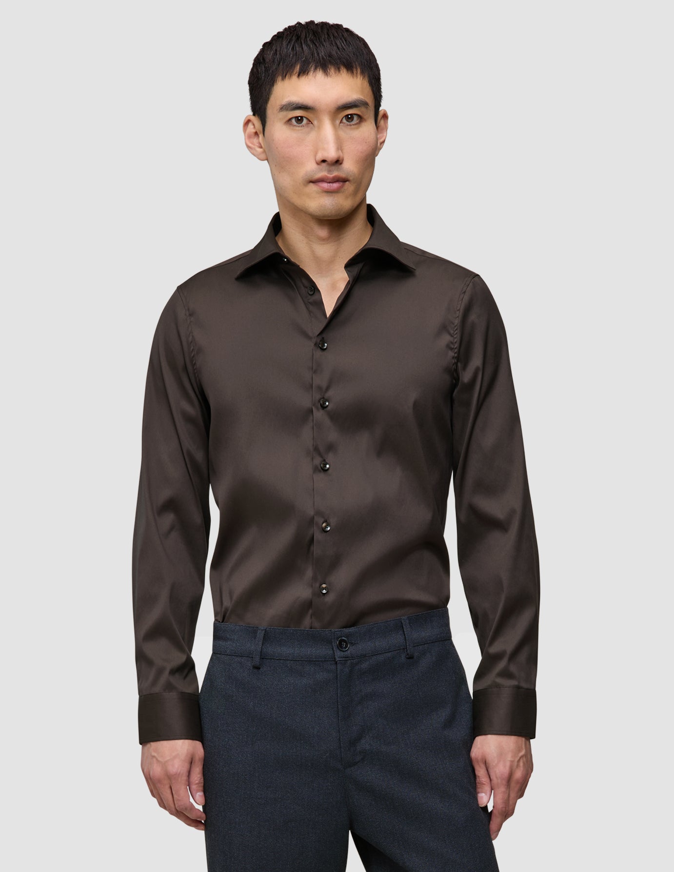 Ein Mann, der ein dunkelbraunes Elevated Shirt Regular trägt, das in eine graue Hose gesteckt ist und ein klares, skandinavisches Design widerspiegelt.