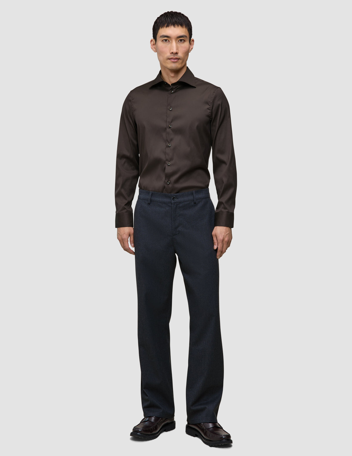 Ein Mann, der ein Elevated Shirt Regular in Dunkelbraun aus Pima-Baumwolle trägt, das in eine anthrazitfarbene Hose gesteckt ist, dazu dunkle Slipper.
