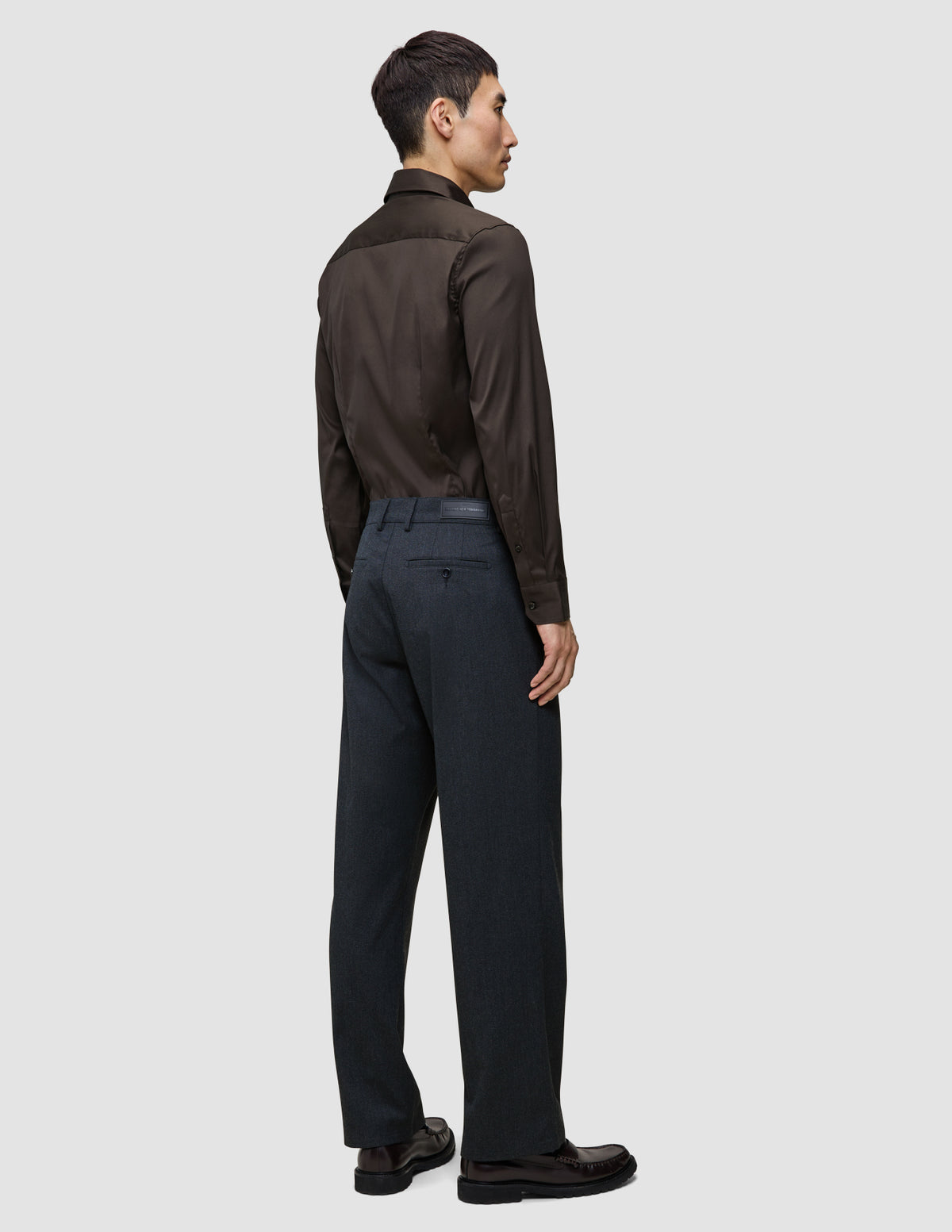 Eine Person, von hinten gesehen, die ein langärmeliges, dunkelbraunes Elevated Shirt Regular-Hemd trägt, das in eine dunkelgraue Hose gesteckt ist, sowie dunkle Leder-Loafer.